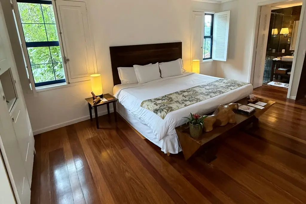 Quarto do Santa Teresa MGallery com cama de casal, piso de madeira e janelas de madeira abertas para o verde
